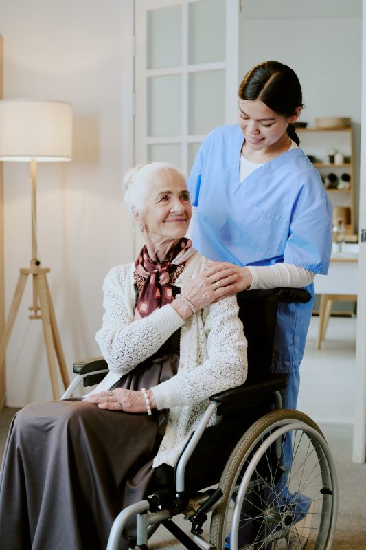 Senior Caucasian woman sitting in wheelchair with disability, smiling while young adult Hispanic female nurse standing behind her, gently placing hands on shoulders, both interacting indoors